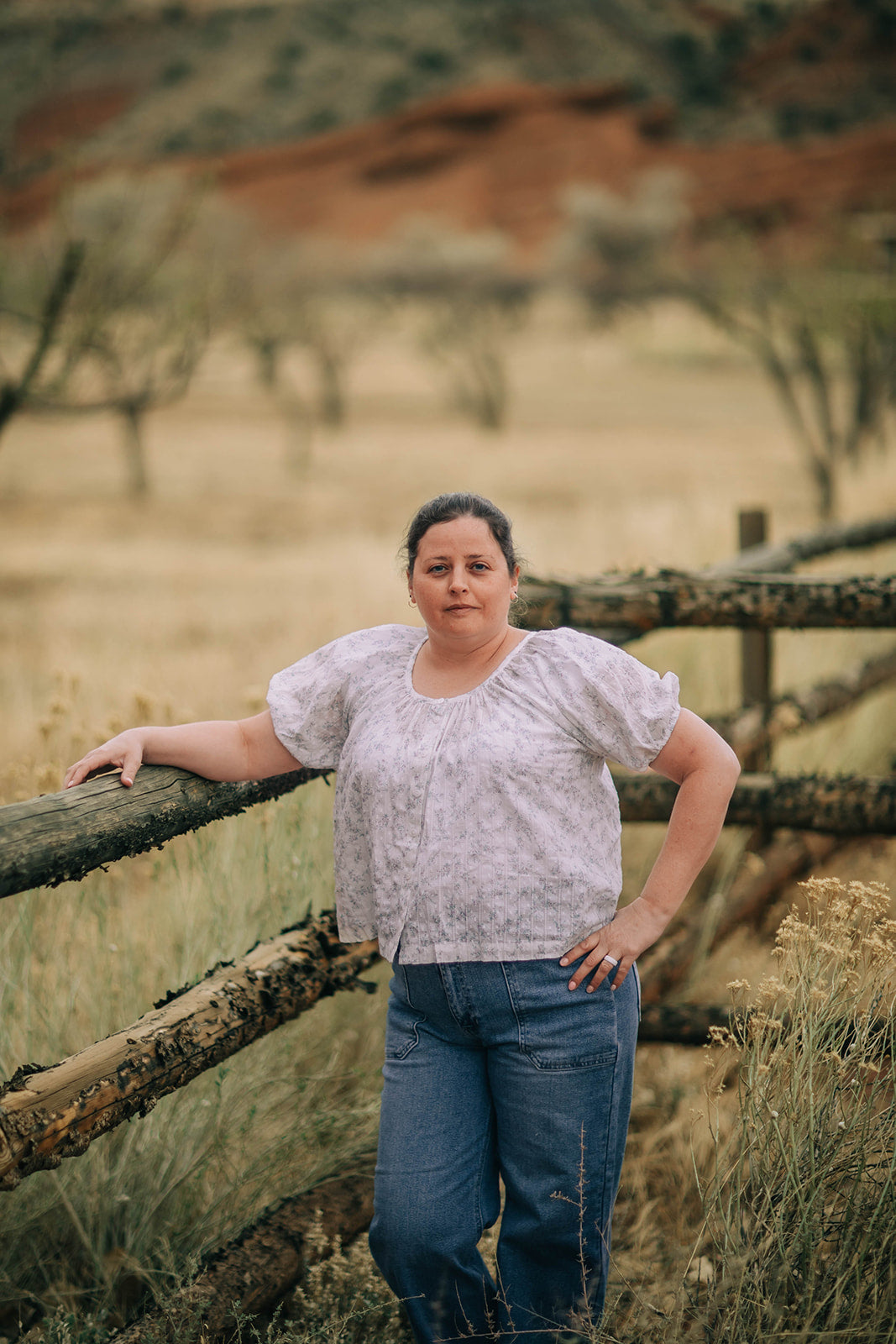 Female model stands in field wearing a button down, floral print, short sleeve, blouse and medium wash jeans.